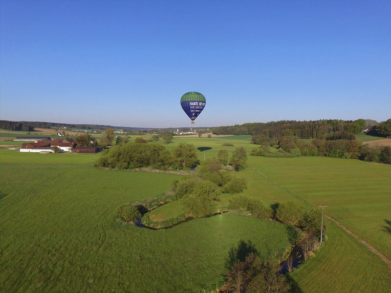 Heißluftballon schwebt über grünen Wiesen einen Flußlauf entlang